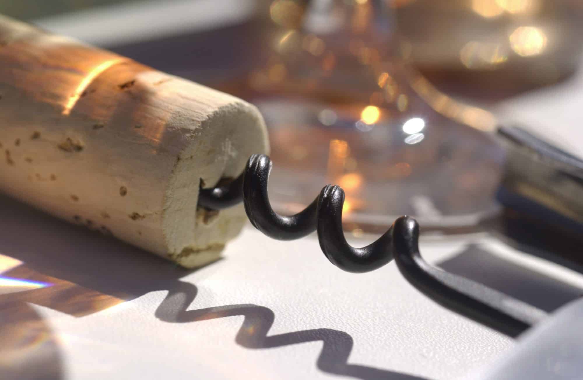 Ein Korken mit Korkenzieher liegt auf einem Tisch vor Weingläsern in denen sich das Licht spiegelt