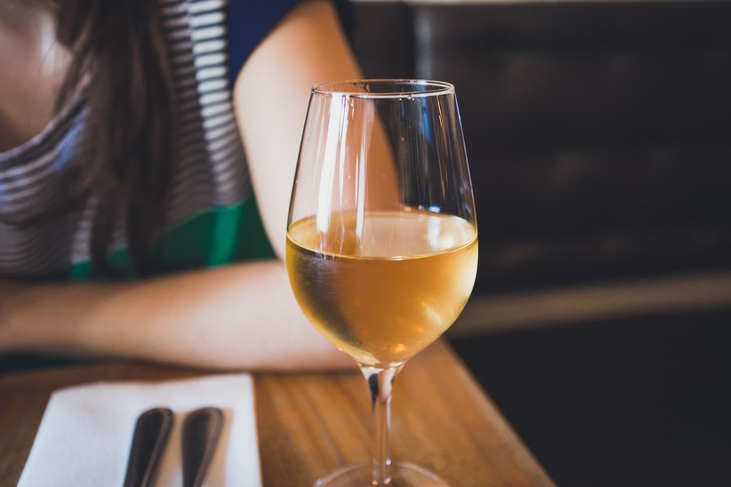 Ein Glas Wein steht auf einem Tisch, eine Person sitzt im Hintergrund